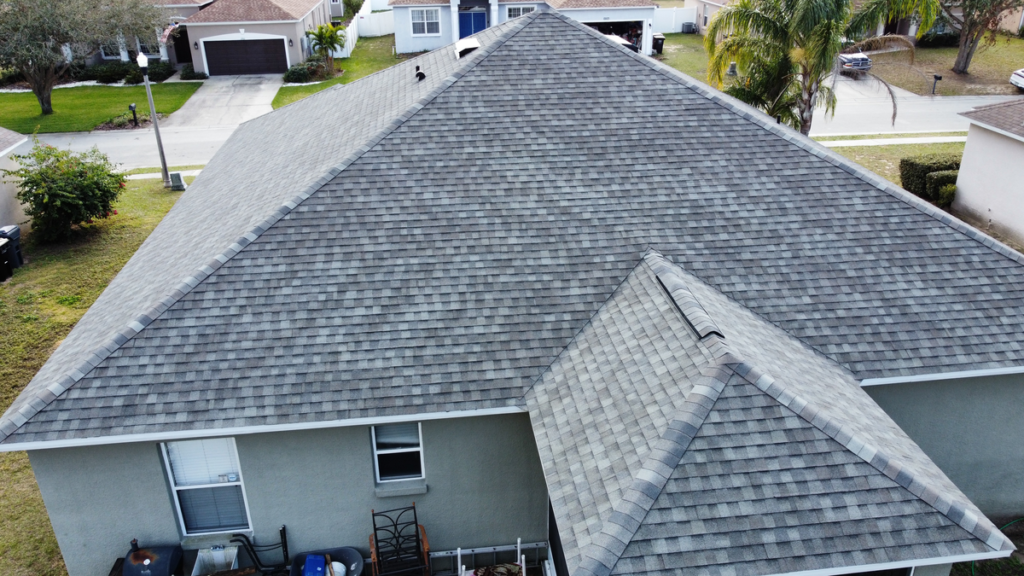 Aerial view of a house with a large, gray shingle roof installed by roofing Wesley Chapel. The backyard features outdoor furniture and storage items near the wall, with neighboring houses, trees, and cars visible along the street in the background.