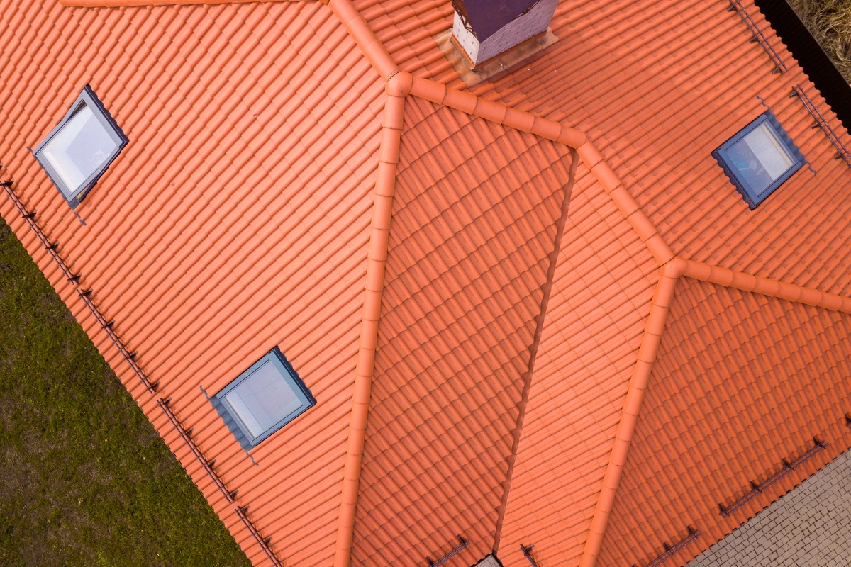 Aerial view of an orange metal Spanish tile roof—known for its durable style and affordable metal Spanish tile roof cost—with four skylight windows, a chimney, green grass, and partial pavement surrounding it.