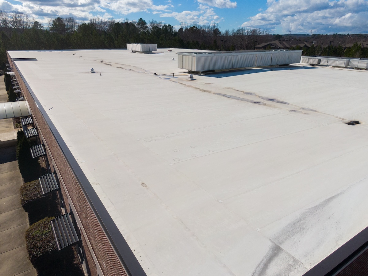 A large, flat, white commercial building roof—possibly showcasing tpo vs epdm materials—features several rooftop units and vents under a partly cloudy sky, surrounded by trees and other buildings in the distance.