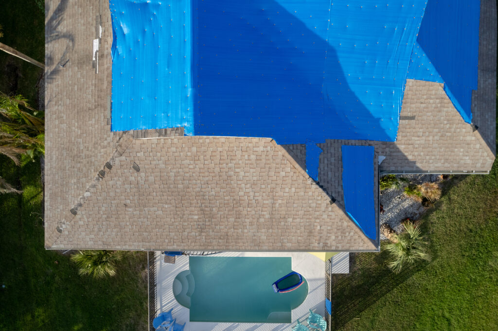 Aerial view of a house with part of its roof covered by an emergency roof tarp, likely for repairs. The backyard features a swimming pool with a floating lounger and shaded patio area; green lawn and palm trees surround the property.