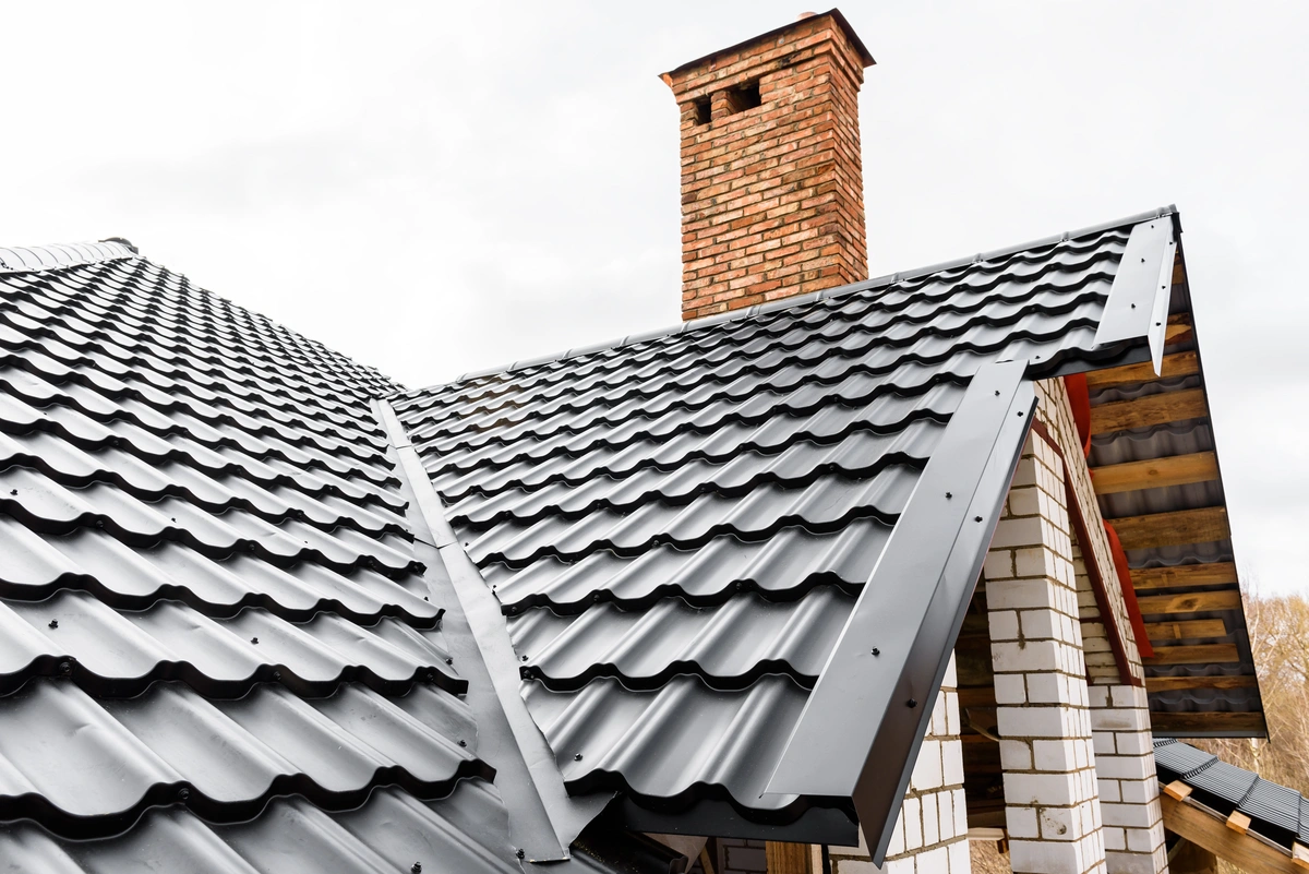 Close-up view of a house roof with black metal roofing tiles and striking metal roof accents, a brick chimney, and white brick walls under a cloudy sky. The roof features a modern design with overlapping sheets and visible fasteners.