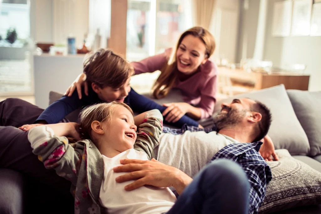 A happy family of four relaxes and laughs together on a couch in a bright living room, feeling joyful and playful under the protection of quality roofing Wesley Chapel residents trust.