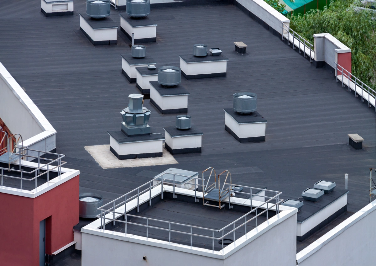 A flat built up roof with multiple square and round ventilation units, small skylights, and safety railings, surrounded by white and red building walls with some greenery visible in the background.