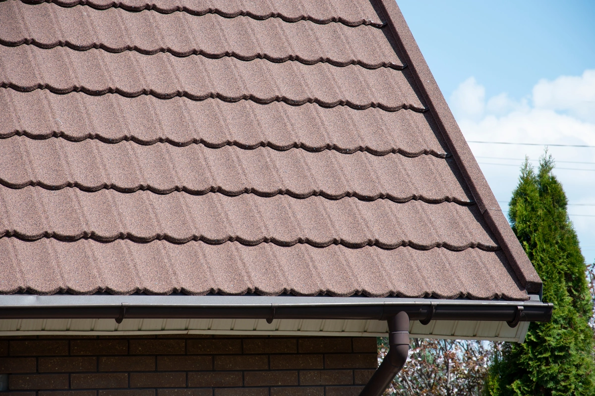 Close-up of a house roof featuring brown, wavy-patterned composite roofing shingles and a rain gutter, with part of a brick wall and some greenery visible in the background under a blue sky.