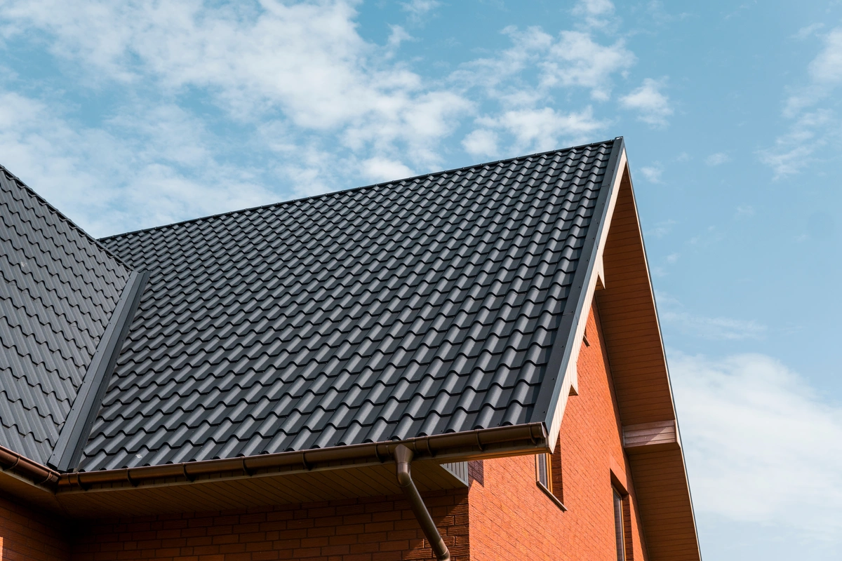 A close-up view of a house with a sloped, dark gray metal roof that looks like shingles, orange brick walls, and a clear blue sky with scattered clouds in the background.