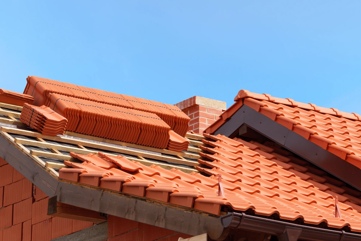 A house roof under construction with stacks of ceramic roof tiles on wooden battens. Part of the roof is already covered with tiles, and a brick chimney is visible against a clear blue sky.