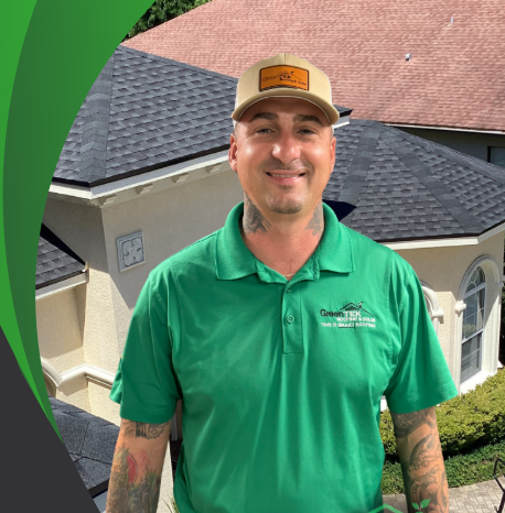 A smiling man in a green polo shirt and tan cap stands in front of a house with a dark shingled roof. He has tattoos on his arms. The background features greenery and part of a two-story home.