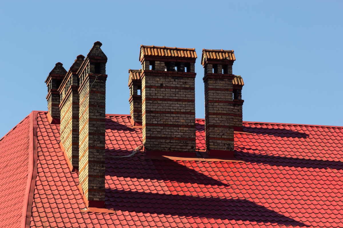 Multiple brick chimneys with decorative tops rise from a red tiled roof, where metal roof flashing neatly seals their bases, casting sharp shadows in the bright sunlight against a clear blue sky.