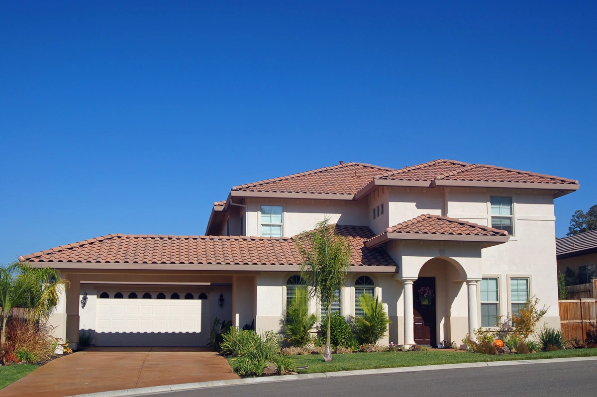 Two-story suburban house with a red-tiled roof under warranty, cream-colored walls, attached garage, and a landscaped front yard with palm trees, all beneath a clear blue sky.