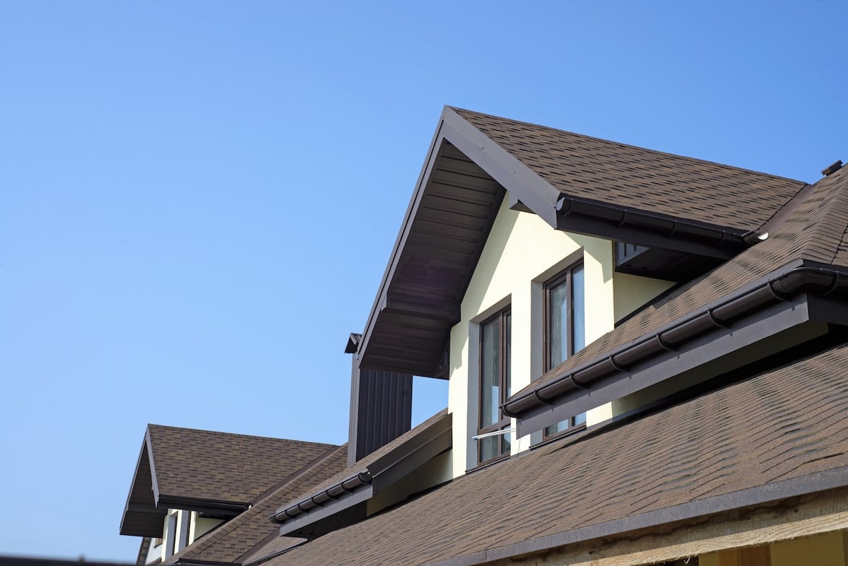 Close-up view of modern house rooftops with brown shingles, triangular gables, and large windows under a clear blue sky, showcasing expert roofing Land O Lakes craftsmanship.