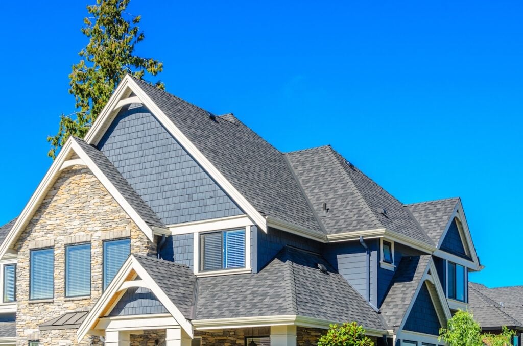 Close-up view of the upper part of a modern house with dark gray shingles by roofing Largo, stone accents, blue siding, and white trim, set against a clear blue sky with a tall tree in the background.