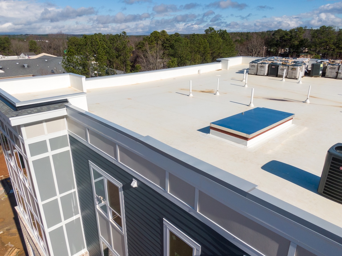 A flat white rooftop with several vents, HVAC units, and a skylight—possibly featuring TPO vs PVC roofing—viewed from above. The building has blue siding and white trim, surrounded by trees under a partly cloudy sky.