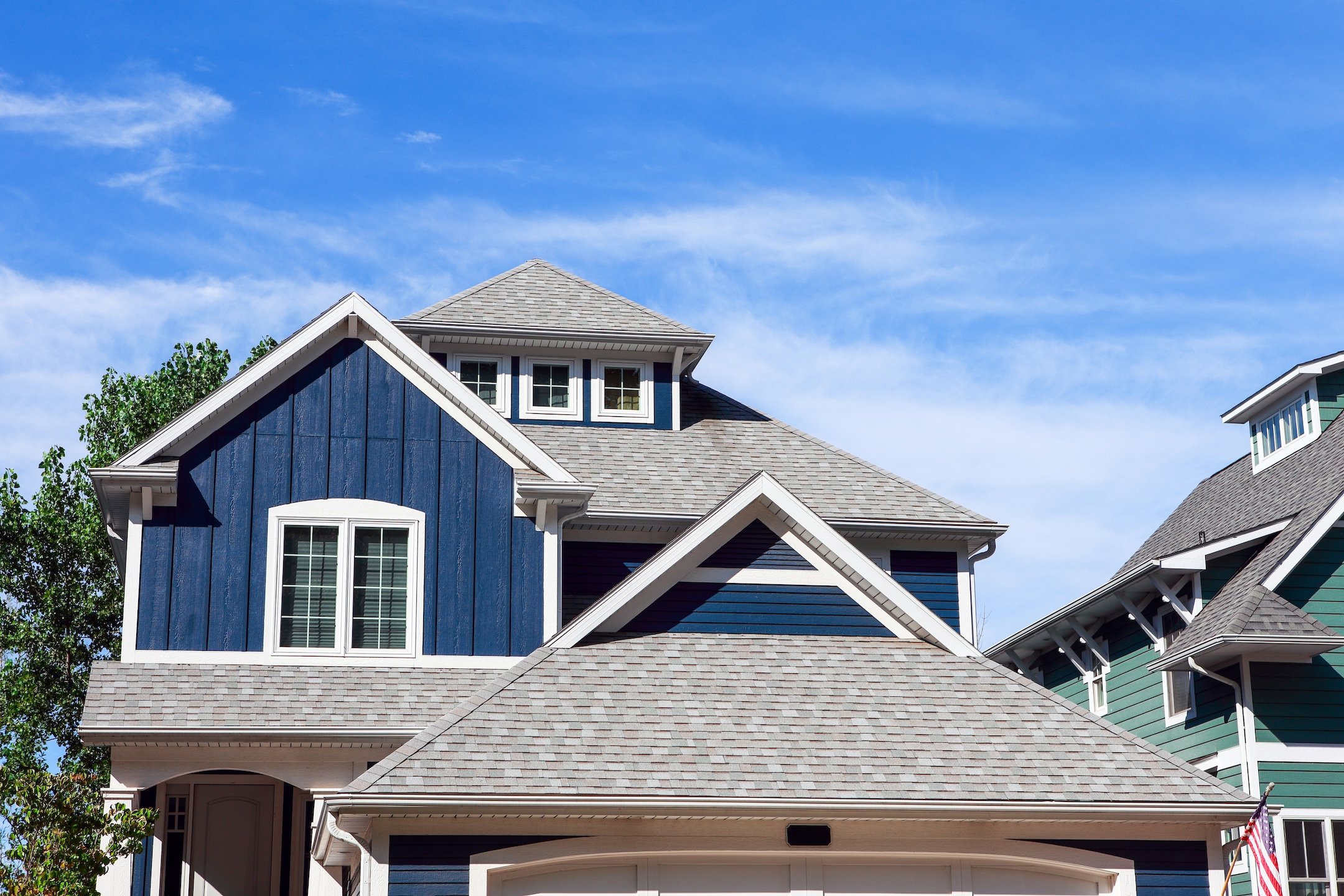 A modern house with blue siding, white trim, and gray shingles showcases expert roofing palm harbor craftsmanship, set against a bright blue sky with wispy clouds. Part of another green house is visible on the right.