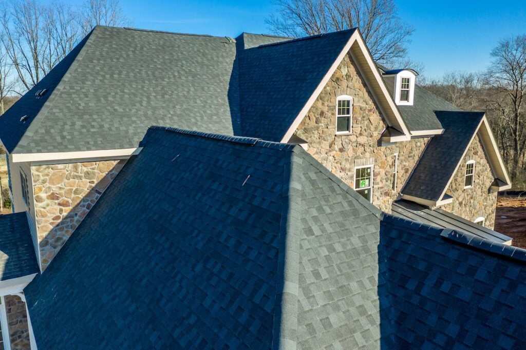 A close-up aerial view of a house with multiple gabled roofs covered in dark gray shingles, expertly installed by roofing Lutz specialists, and stone exterior walls with several windows, surrounded by leafless trees under a clear blue sky.