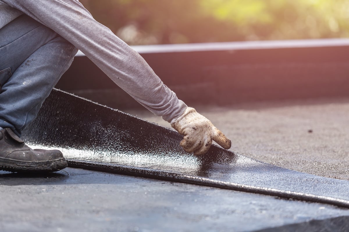 A person wearing gloves installs a roof membrane on a flat roof, carefully smoothing it out with their hand. The freshly laid surface catches the sunlight in the background.