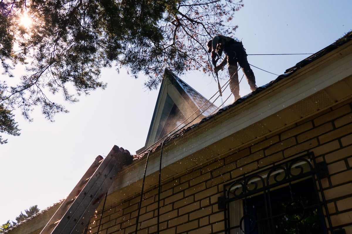 A person in safety gear performs roof pressure washing, standing near the peak of a steep house roof as water sprays. A tall ladder is propped against the brick building, with sunlight filtering through trees overhead.