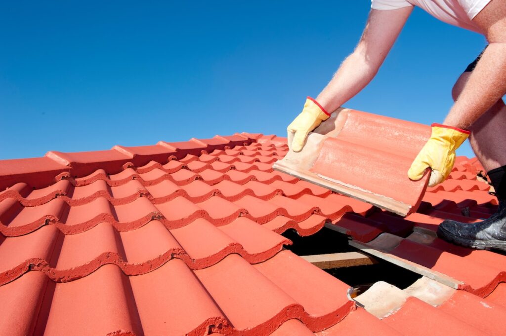 Roof repair, worker with yellow gloves replacing red tiles or shingles on house with blue sky as background and copy space. tile roof leak repair