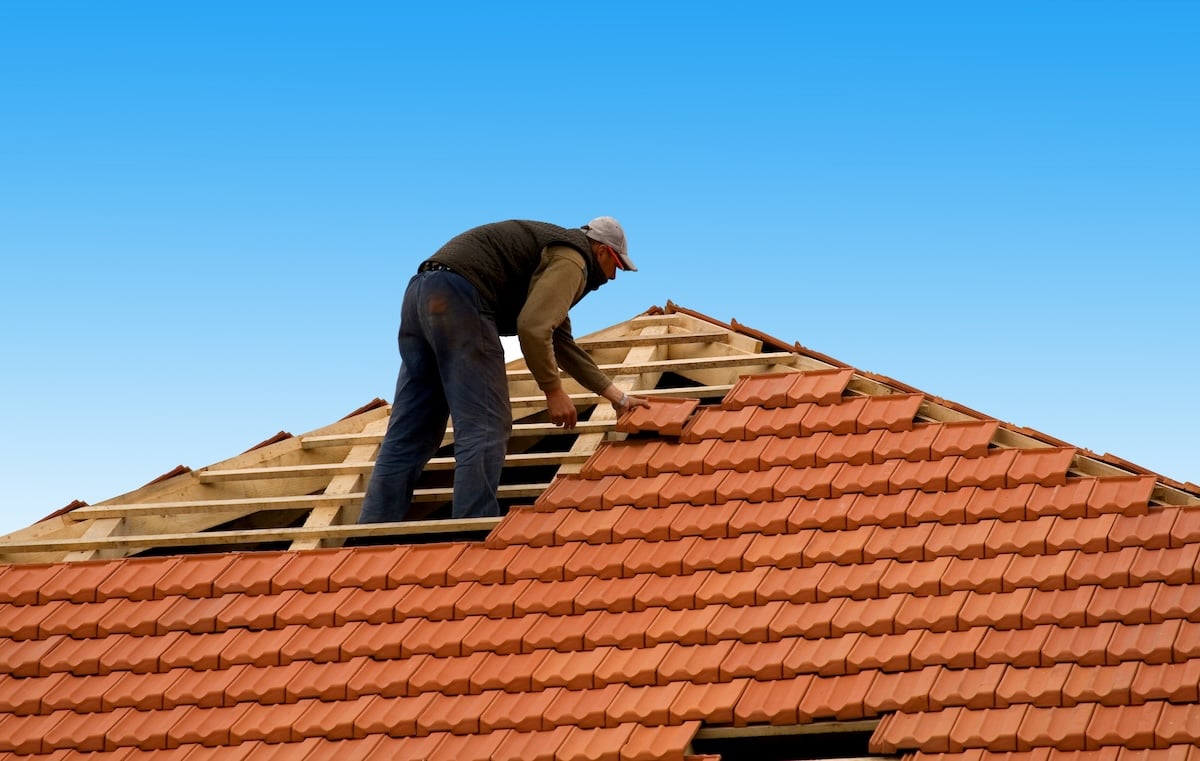 A person works on tile roof leak repair, installing or fixing red roof tiles atop a house while standing on the wooden framework under a clear blue sky.