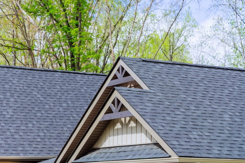The image shows the roof of a house with dark gray shingles and decorative wooden trim, highlighting expert roofing Lutz craftsmanship, set against tall trees with green leaves and a blue sky.