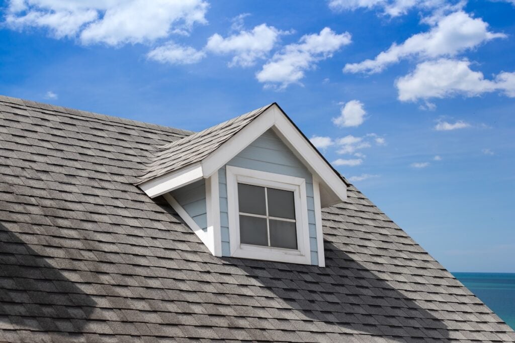 A close-up of a house roof with gray shingles installed by Roofing Ruskin, featuring a small dormer window. The sky is bright and blue with scattered white clouds, and the ocean is visible in the background.