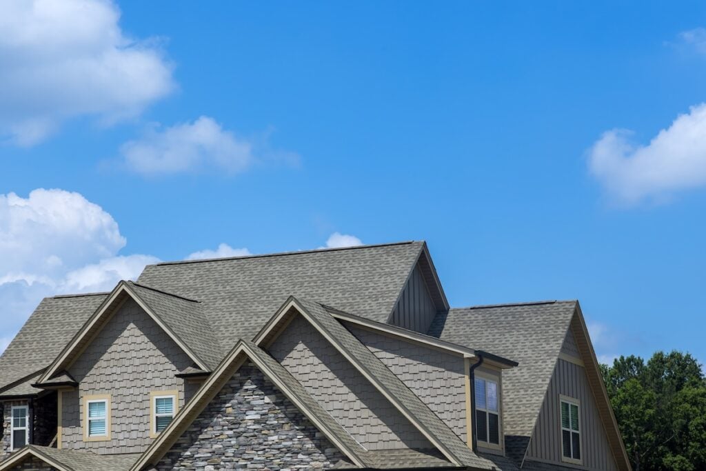 The image shows the upper portion of a modern house with gray siding and stone accents, set against a bright blue sky with scattered white clouds. Trees are visible in the background, highlighting expert roofing Palm Harbor craftsmanship.
