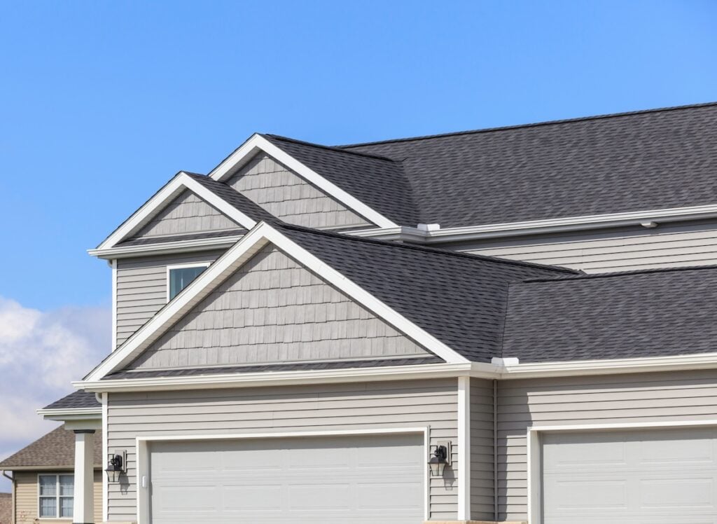 Close-up of a modern suburban house exterior featuring light gray siding, shingle accents, white trim, and double garage doors—showcasing premium roofing Treasure Island style—set against a clear blue sky.