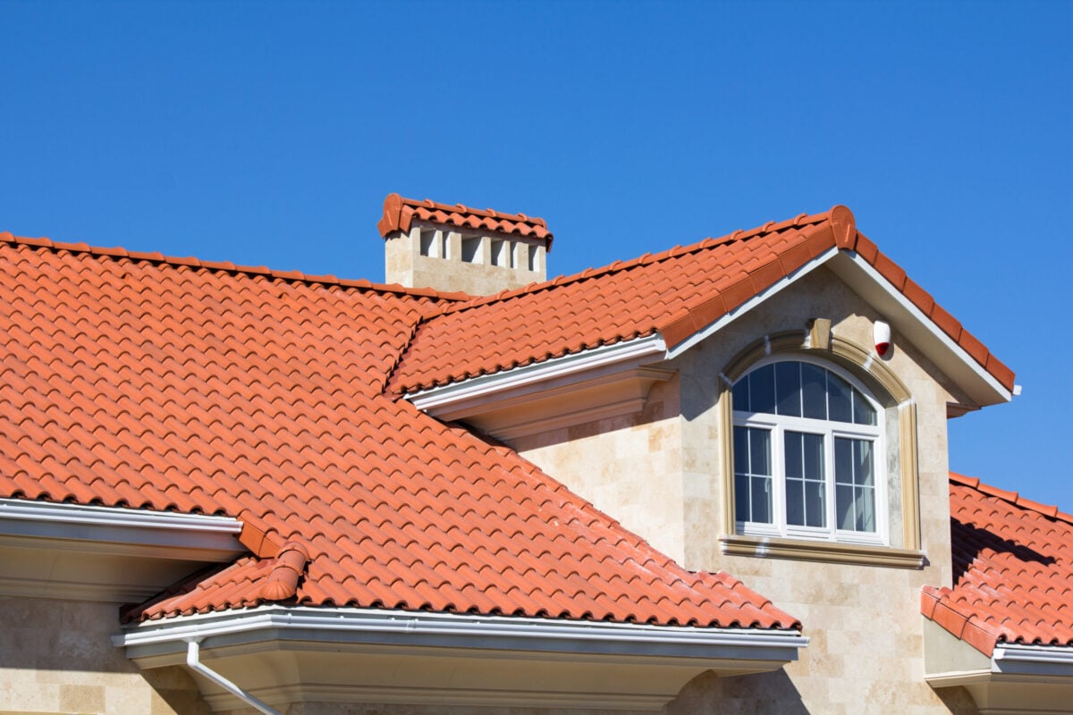 House with beige stone exterior and red clay tile roof, featuring a dormer window with an arched top and white trim, under a clear blue sky—a perfect example for those learning how to install clay roof tile.