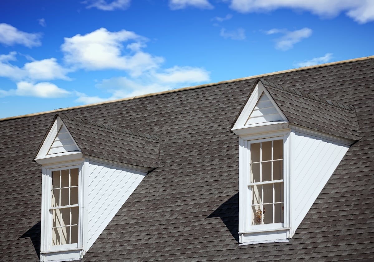 A close-up of a roof with gray shingles featuring two white dormer windows under a bright blue sky with scattered clouds, perfect for showcasing new roof financing options.