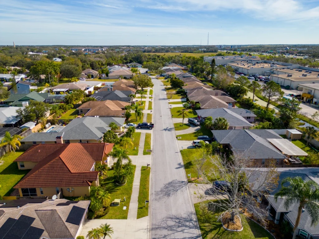 Aerial view of a suburban neighborhood with rows of single-family houses, green lawns, and driveways lining both sides of a straight, quiet street under a partly cloudy sky.