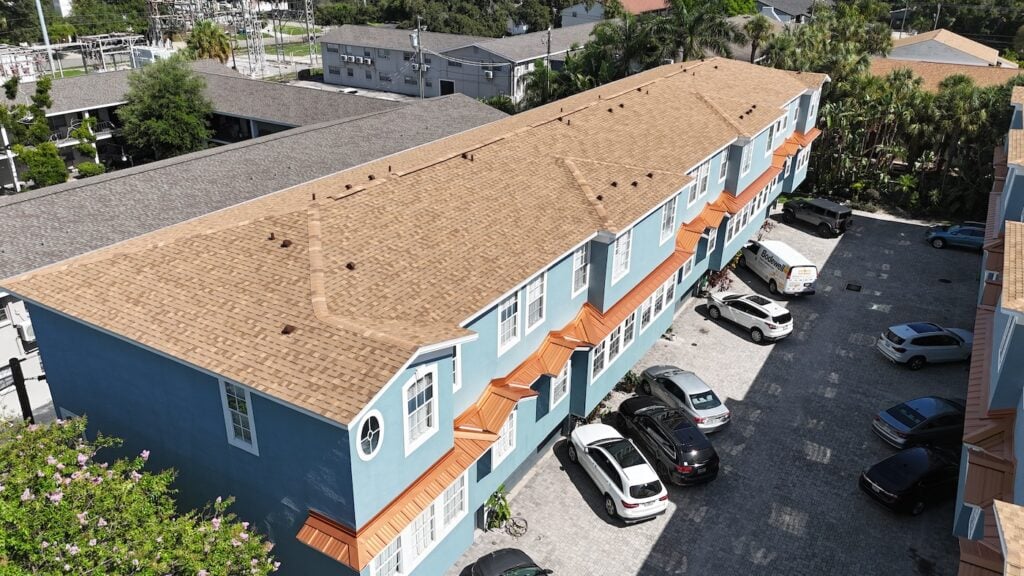Aerial view of a blue two-story apartment building with a tan roof, orange awnings, and multiple parked cars in a paved parking lot beside the building on a sunny day.