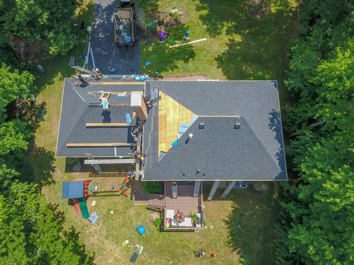 Aerial view of a house with workers repairing the roof; shingles and materials are spread out. A crane truck is parked on the driveway, and the yard is surrounded by trees with scattered tools and play equipment visible.