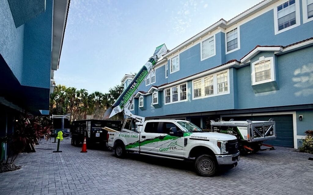 A white Geotex Solutions truck with equipment is parked in a paved driveway between blue multi-story homes, with a worker and trees visible in the background.