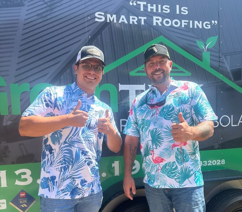 Two men wearing tropical-patterned shirts and baseball caps stand smiling in front of a vehicle with roofing advertisement graphics. Both are giving thumbs up and appear cheerful and relaxed.