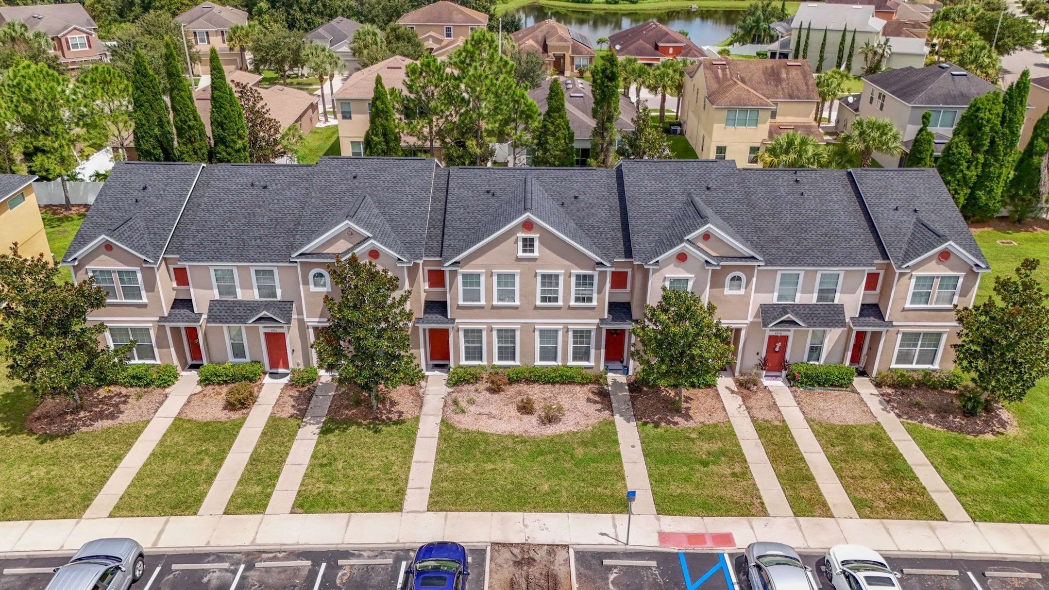 Aerial view of a row of connected two-story townhouses with red doors, small front lawns, and trees, situated in a suburban neighborhood with parked cars in front.