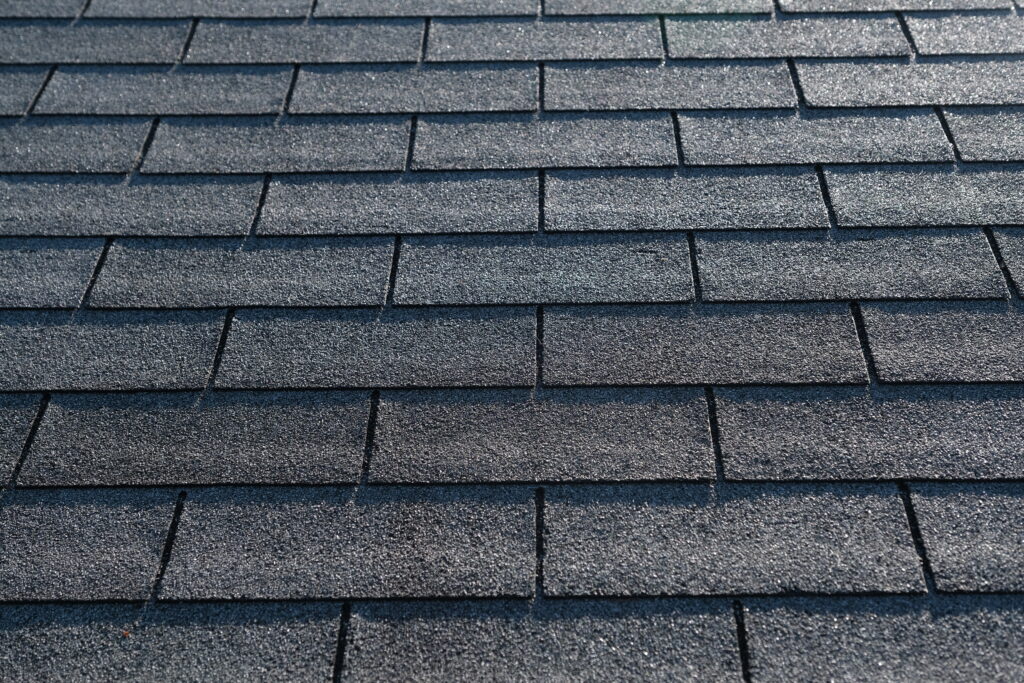 Close-up view of a roof covered with overlapping rectangular composition shingles, arranged in a neat, uniform pattern. The shingles have a rough, textured surface in varying shades of gray.
