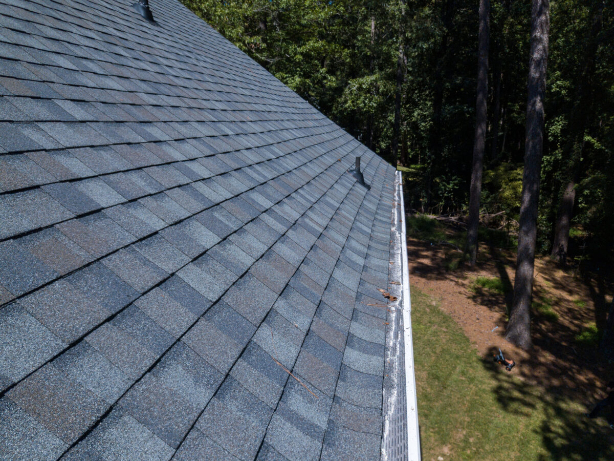 A close-up view of a house roof with gray composition shingles and a white gutter, surrounded by tall trees and a grassy yard below.