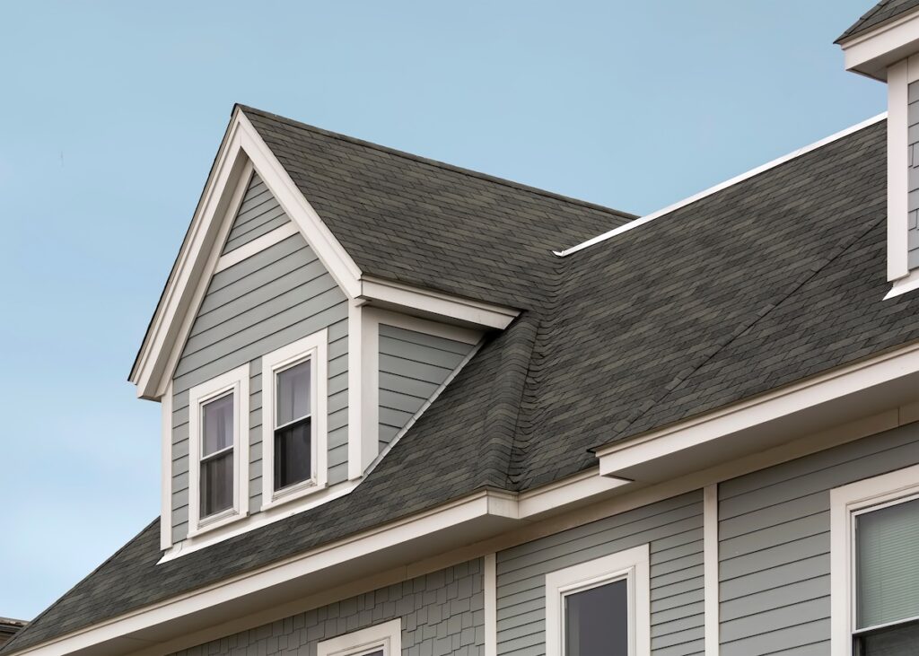 A close-up view of a house with gray siding and white trim, featuring a gable roof with dark composition shingles and double-hung windows, set against a clear blue sky.