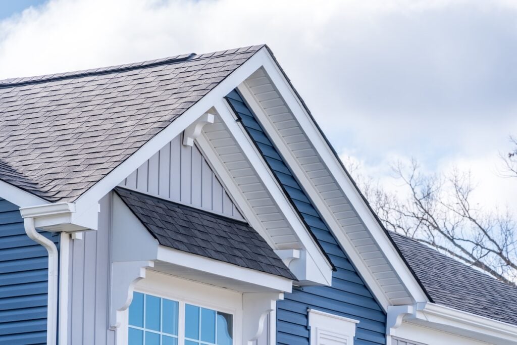 Close-up of the upper section of a blue house with white trim, showing gable rooflines, decorative brackets, and large windows beneath a partly cloudy sky, featuring Tamko shingle colors on the roof.