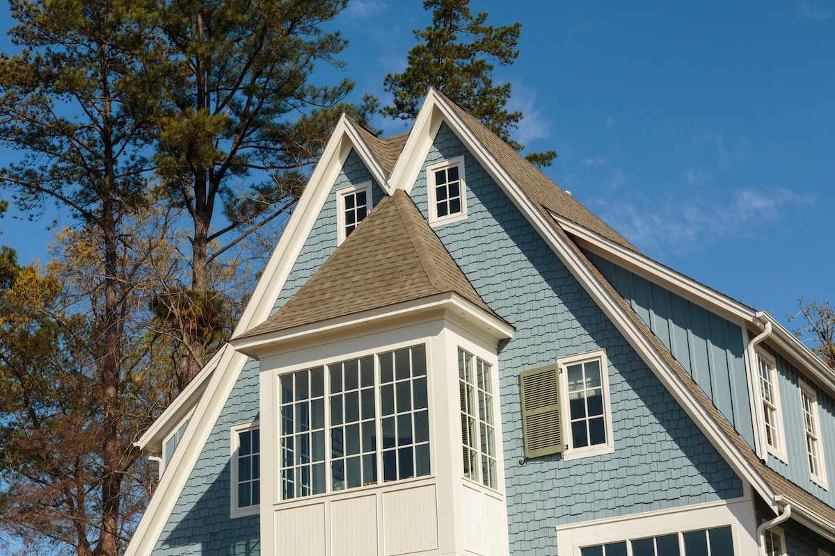 The image shows the upper part of a blue house with white trim, large windows, and multiple steep gables—ideal for comparing tamko vs gaf shingles. Tall pine trees stand in the background under a clear blue sky.
