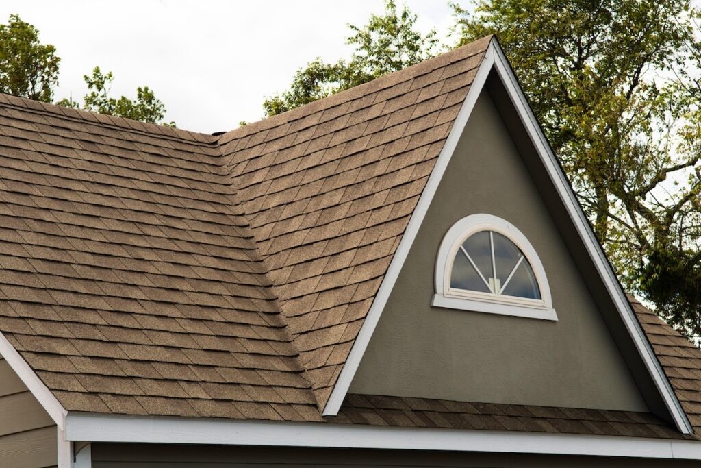 A close-up of a house roof with brown Tamko shingle colors and a gable. The gable features a small, white-trimmed arched window. Green trees are visible in the background under a cloudy sky.