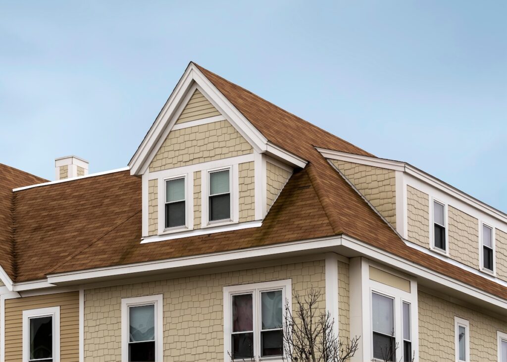 A beige house with tan shingles and white trim, featuring a steeply pitched roof, dormer windows, and an overcast sky—perfect for showcasing tamko vs gaf shingles in terms of style and durability. tamko vs gaf shingles