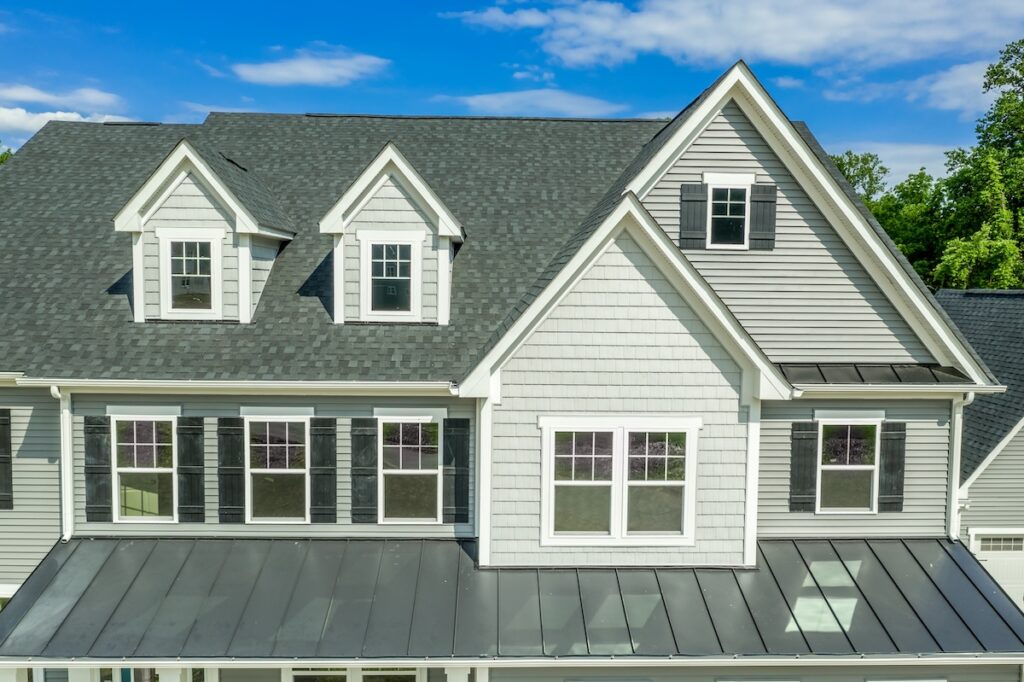 Close-up view of the upper front exterior of a light gray house with black shutters, multiple windows, dormers, and a metal roof section under a clear blue sky—perfect for comparing tamko vs gaf shingles. tamko vs gaf shingles