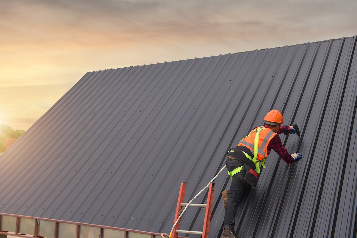 A construction worker wearing a safety helmet, vest, and harness is installing or inspecting a dark metal roof panel while standing on a ladder at sunset.