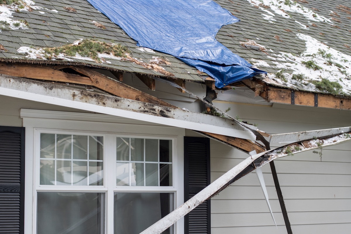 A house with signs of storm damage to the roof is covered by a blue tarp. Exposed wooden beams and insulation are visible, along with broken gutters and some snow and debris scattered on the roof and ground.