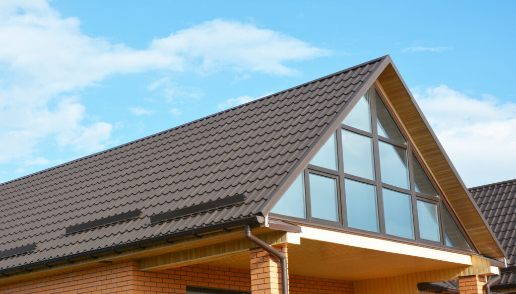 A close-up view of a house roof with brown shingles and large triangular windows, set against a blue sky with scattered clouds. The exterior walls are made of light-colored bricks. how to install metal roofing