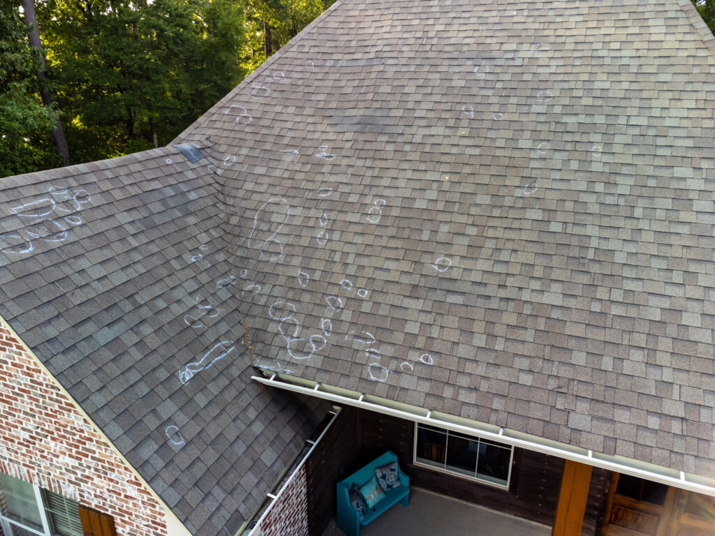 Aerial view of a house roof shows numerous chalk circles and marks indicating possible signs of storm damage to roof shingles. Trees are visible in the background with a patio area seen below. signs of storm damage to roof
