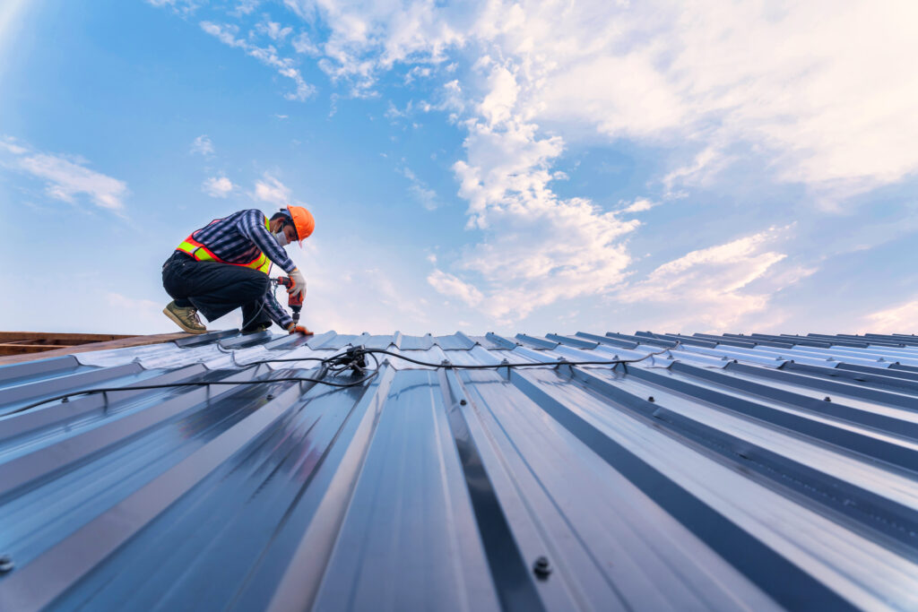 A worker wearing a safety vest and helmet is installing or repairing a metal roof under a blue sky with scattered clouds. The person is using tools and safety equipment while working on the sloped rooftop.