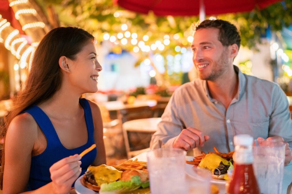 A smiling couple sits at an outdoor restaurant table at night, eating burgers and fries. Decorative string lights and greenery create a warm, festive atmosphere—one of the charming things to do in Wesley Chapel. things to do in wesley chapel