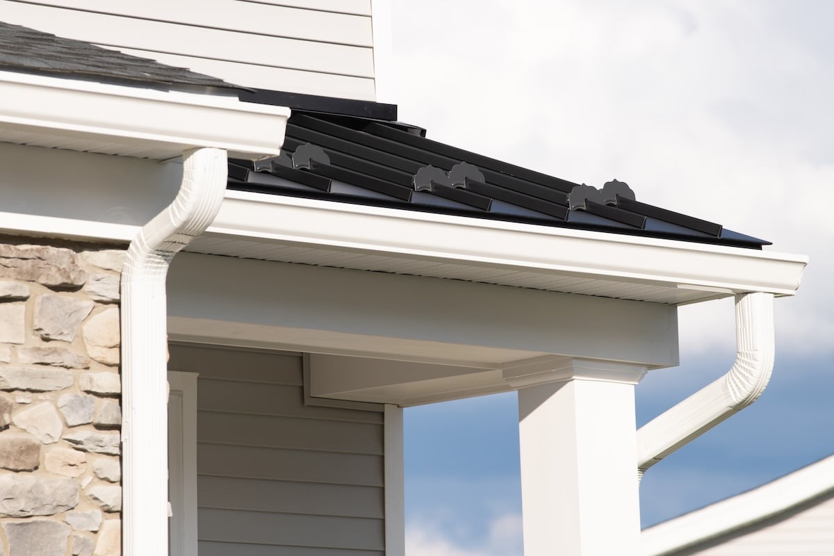 Close-up of a house’s corner featuring sleek white gutters, a metal roof with decorative bat-shaped accents, light gray siding, a stone wall section, and a white column under a partly cloudy sky.