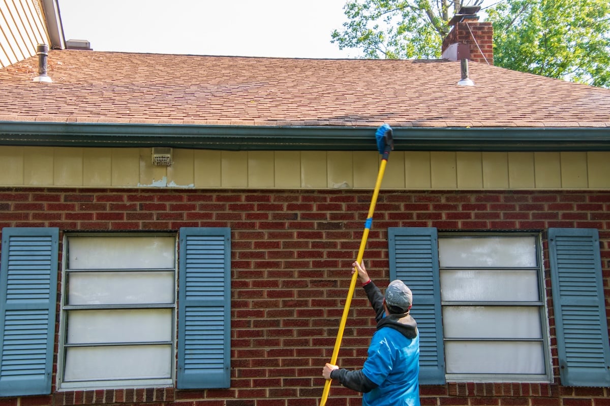 A person uses a long yellow pole with a brush to clean the gutters of a brick house with blue shutters and a brown roof, showing the care and attention often seen in soft wash roof cleaning. soft wash roof cleaning